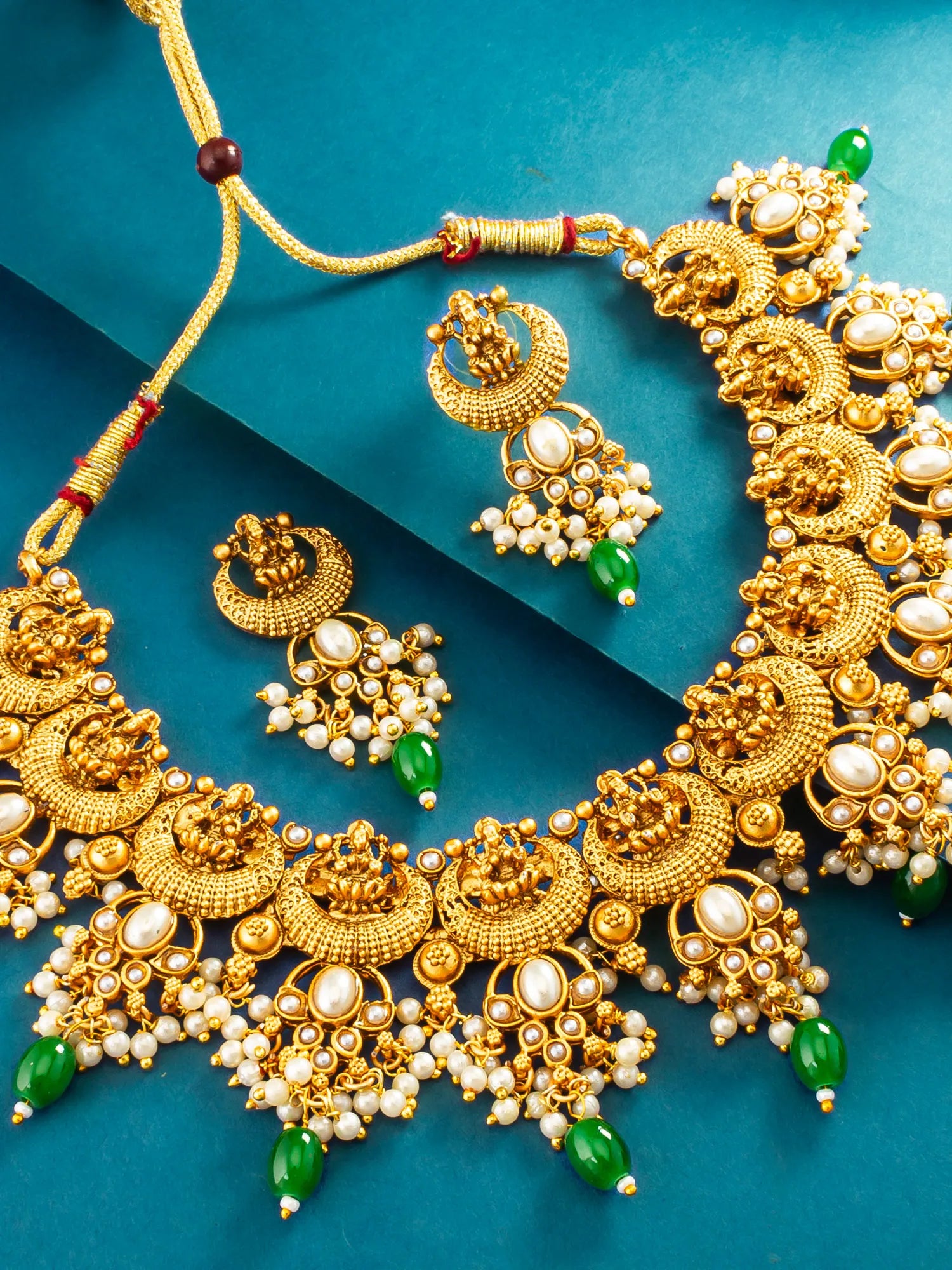 An ornate gold-toned traditional necklace with matching dangle earrings, decorated with white faux pearls, green bead accents, and intricate filigree details, arranged on a teal-blue background.