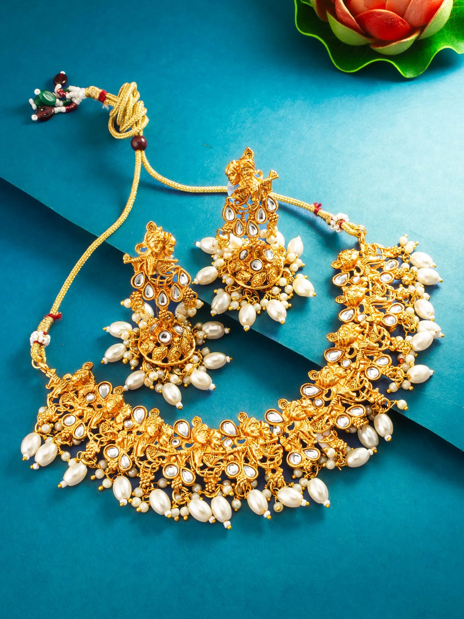 An ornate gold-toned traditional jewelry set with a necklace, earrings, and a maang tikka, decorated with white pearl-like beads and small red and green accents, arranged on a teal background with a decorative flower in the corner.