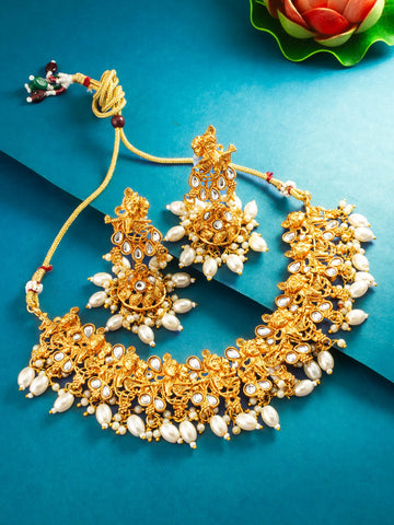 An ornate gold-toned traditional jewelry set with a necklace, earrings, and a maang tikka, decorated with white pearl-like beads and small red and green accents, arranged on a teal background with a decorative flower in the corner.