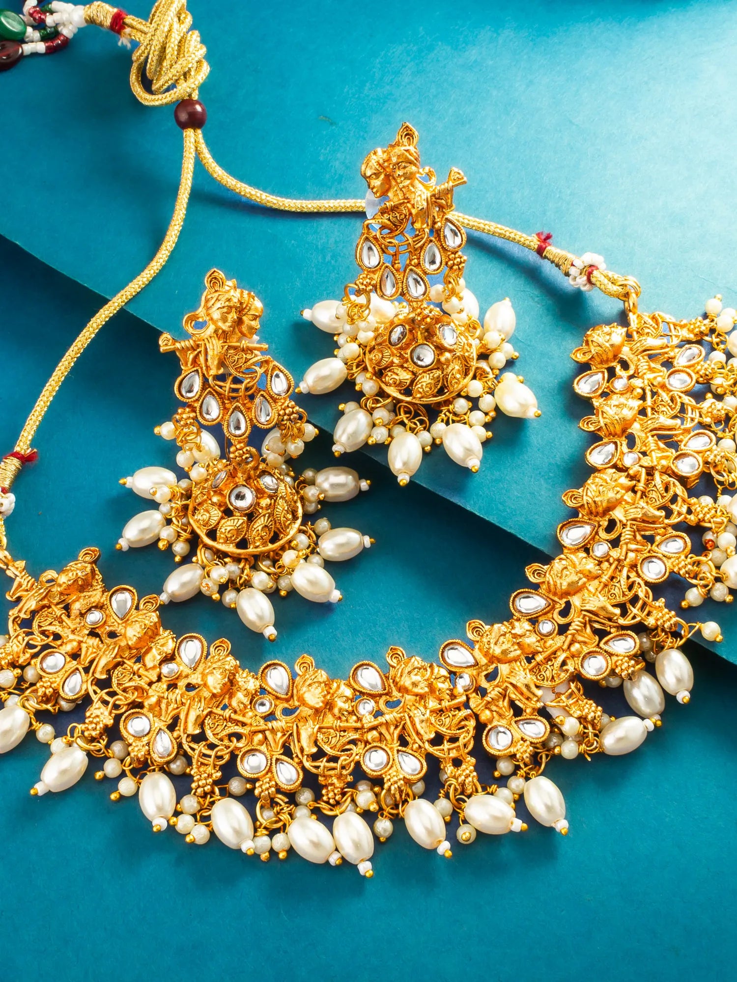 An ornate gold-toned traditional jewelry set with matching earrings, a statement necklace, and pearl-like bead accents, arranged on a teal-blue background.