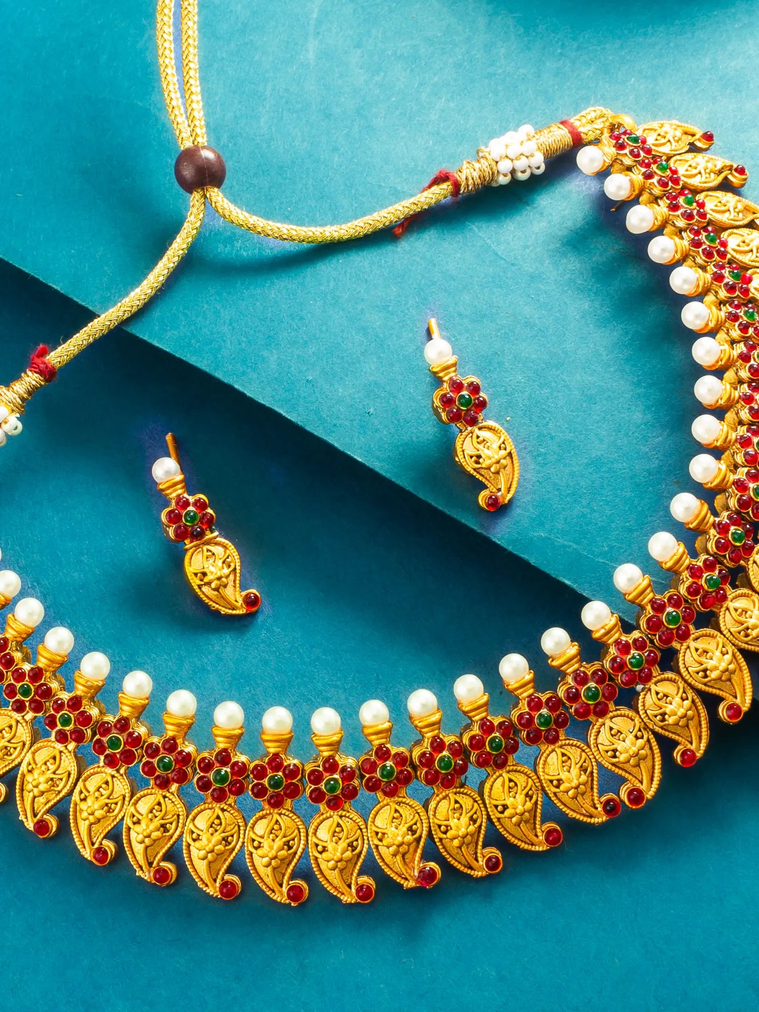 Gold-toned traditional necklace and matching earrings with white pearl-like beads and red and green stone accents, arranged on a teal background.