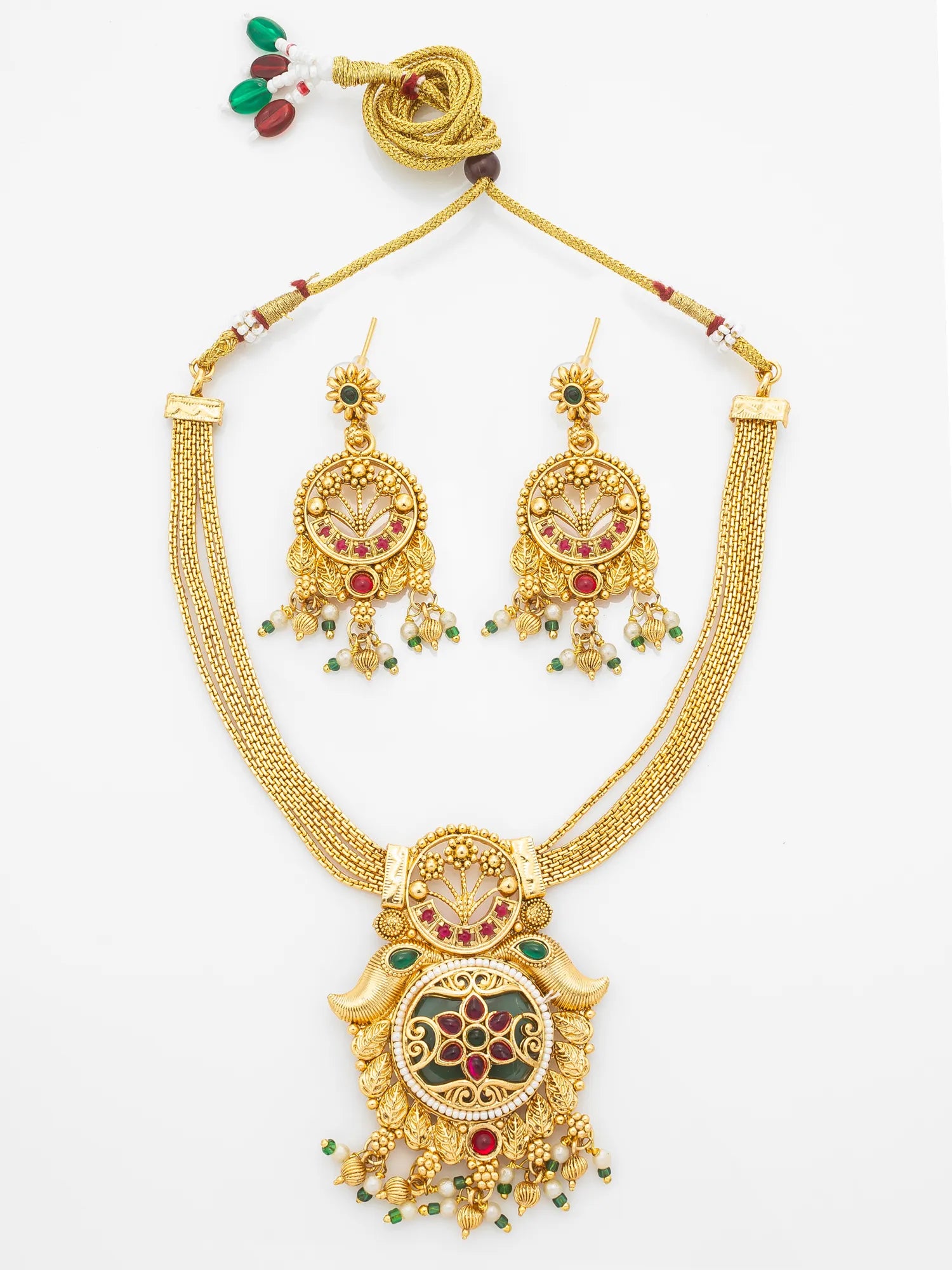 Gold-plated traditional Indian jewelry set featuring a necklace and matching earrings with intricate filigree work, red and green stone accents, and a decorative hanging cord with beads, displayed on a clean white background.