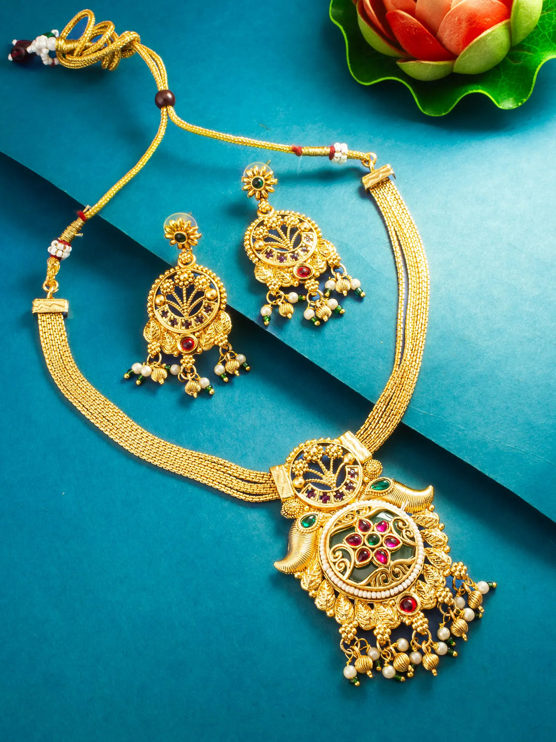 Gold-toned traditional Indian jewelry set featuring a necklace with a large ornate pendant, matching drop earrings, and a beaded cord, arranged on a teal-blue background with decorative floral elements.