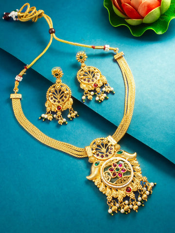 Gold-toned traditional Indian jewelry set featuring a necklace with a large ornate pendant, matching drop earrings, and a beaded cord, arranged on a teal-blue background with decorative floral elements.