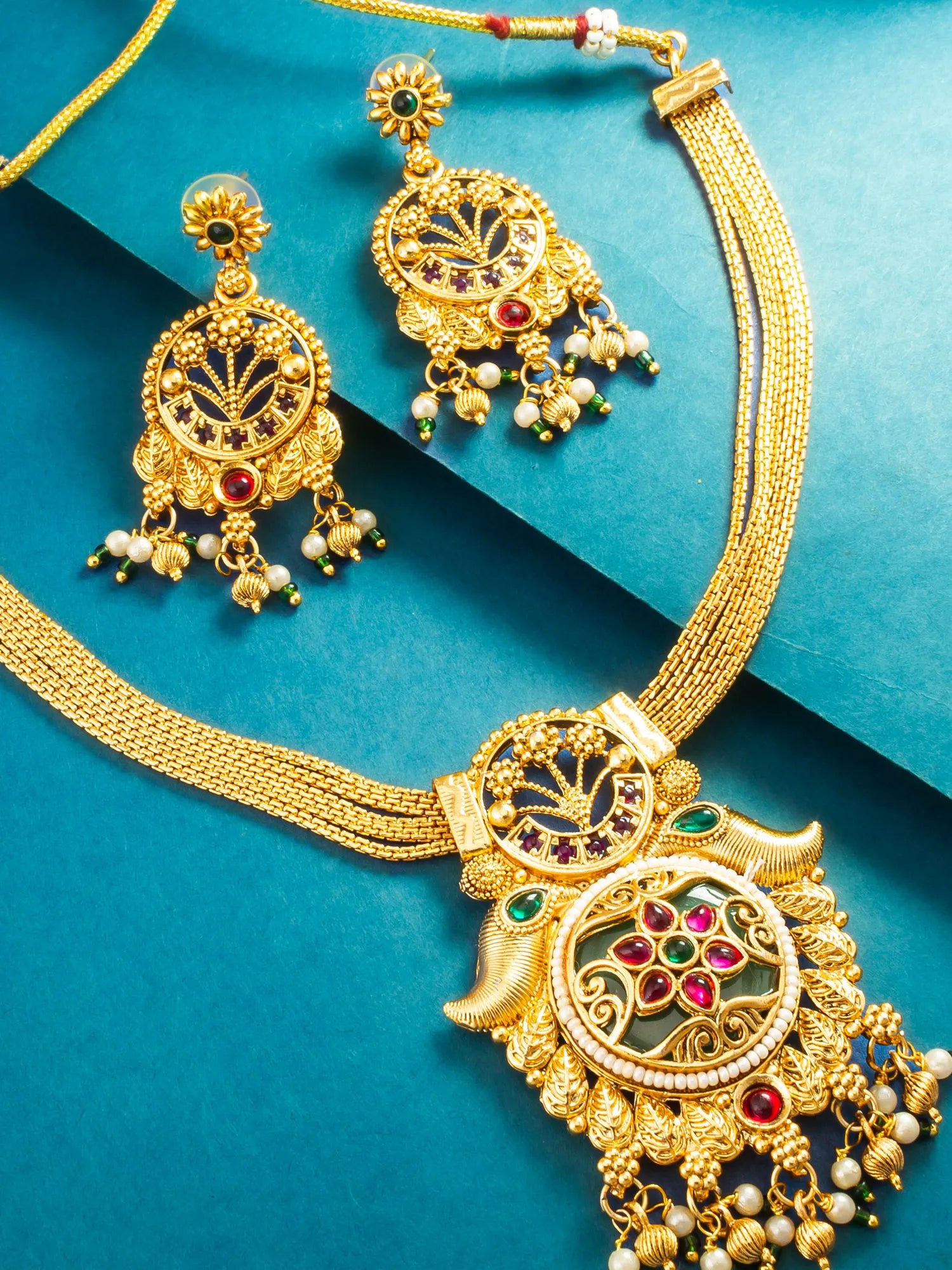Gold-toned traditional jewelry set featuring a necklace and matching earrings with ornate filigree, red and green stone accents, and small pearl-like beads, arranged on a teal background.