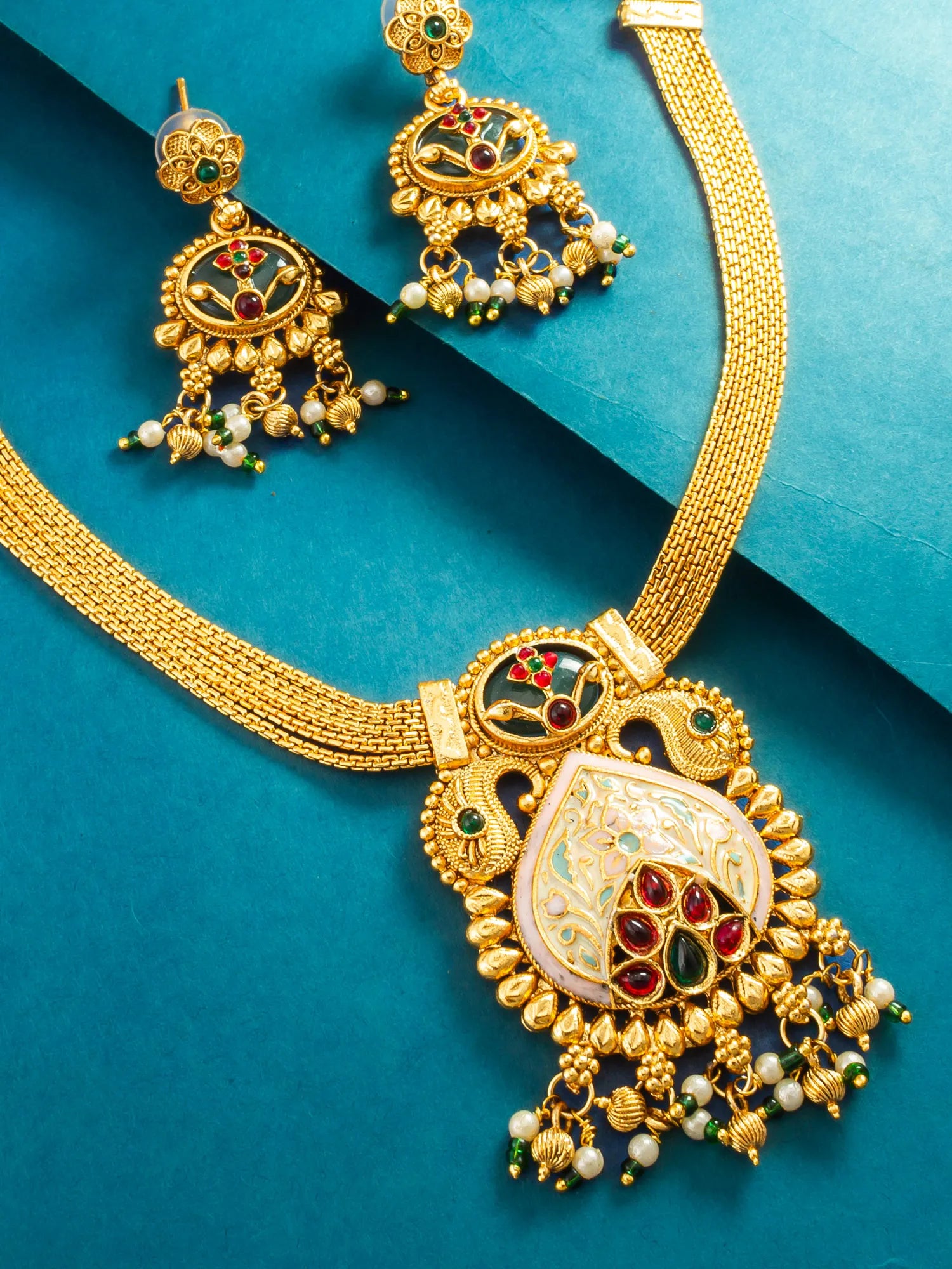 Gold-toned traditional Indian jewelry set featuring a necklace and matching earrings with intricate filigree, red and green enamel details, and white pearl-like beads, displayed on a teal background.