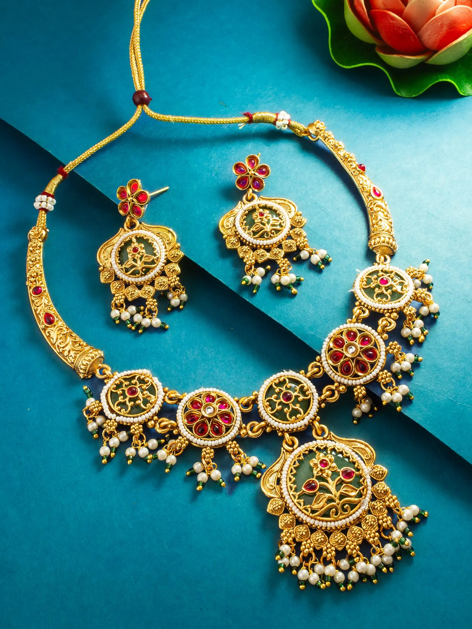 An ornate gold-toned traditional necklace and matching drop earrings with red floral inlays, white pearl bead drops, and green enamel details, arranged on a blue background with a decorative flower in the corner.