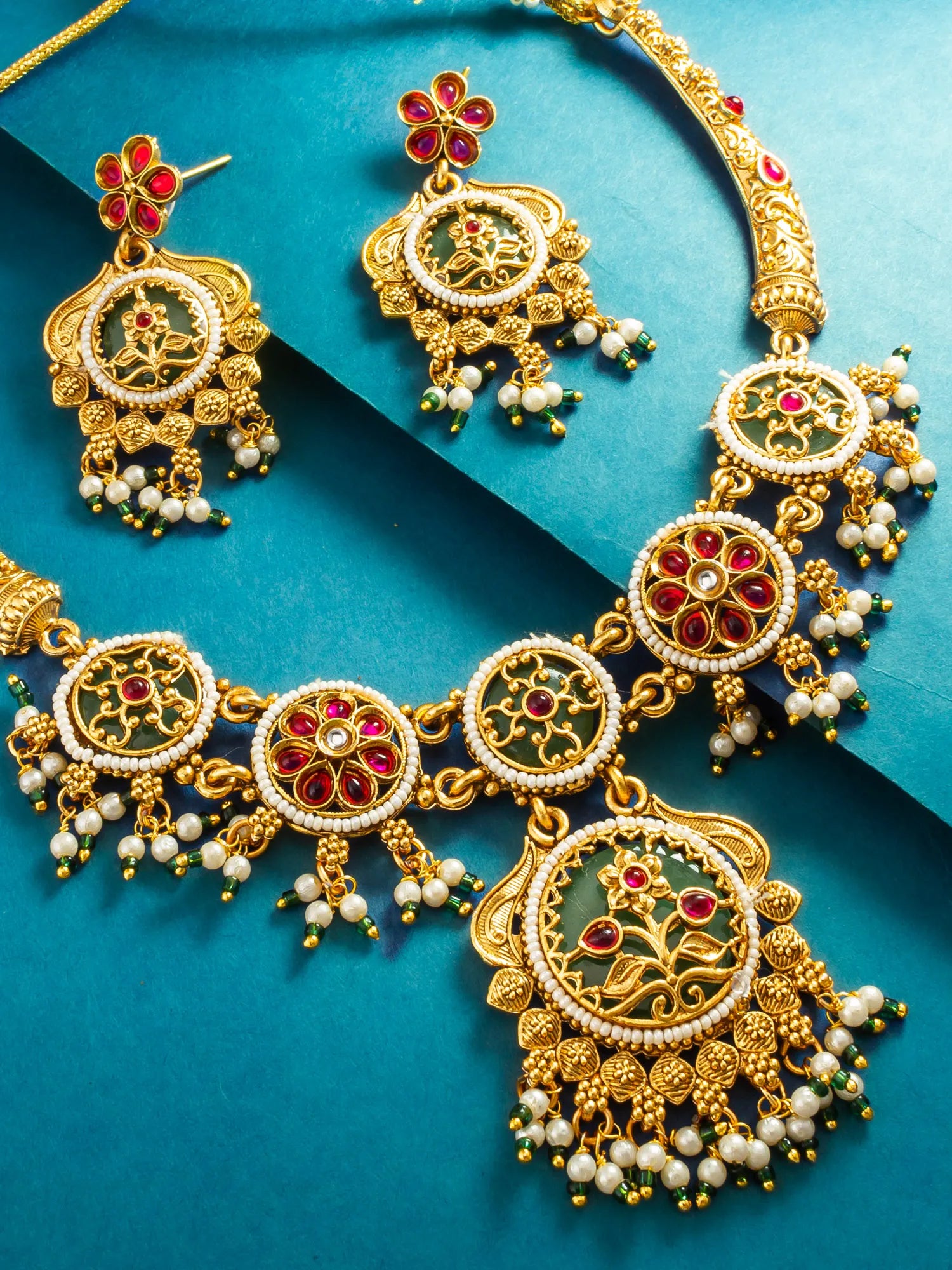 An ornate gold-toned jewelry set with a necklace and matching earrings, featuring circular motifs decorated with white faux pearls, red gemstones, and green enamel details, displayed on a teal background.