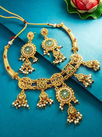 An ornate gold-toned traditional jewelry set including a necklace, matching earrings, and dangling charms, decorated with green stones, red accents, and white pearls, arranged on a blue background with a decorative lotus leaf in the corner.