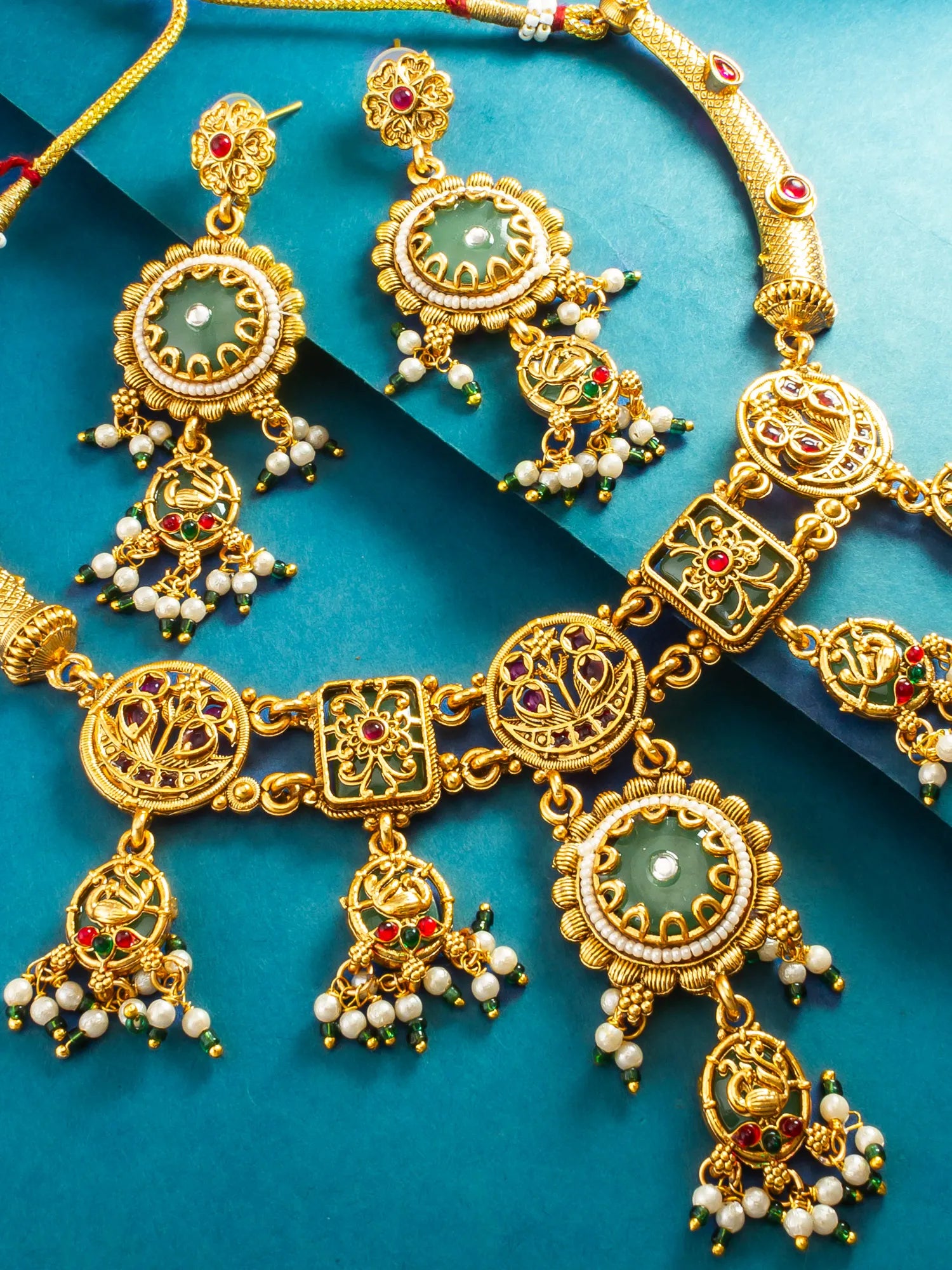 An ornate gold-toned jewelry set with matching necklace, earrings, and pendant, featuring green stone centers, white pearl-like beads, and small red and green jewel accents, arranged on a teal background.