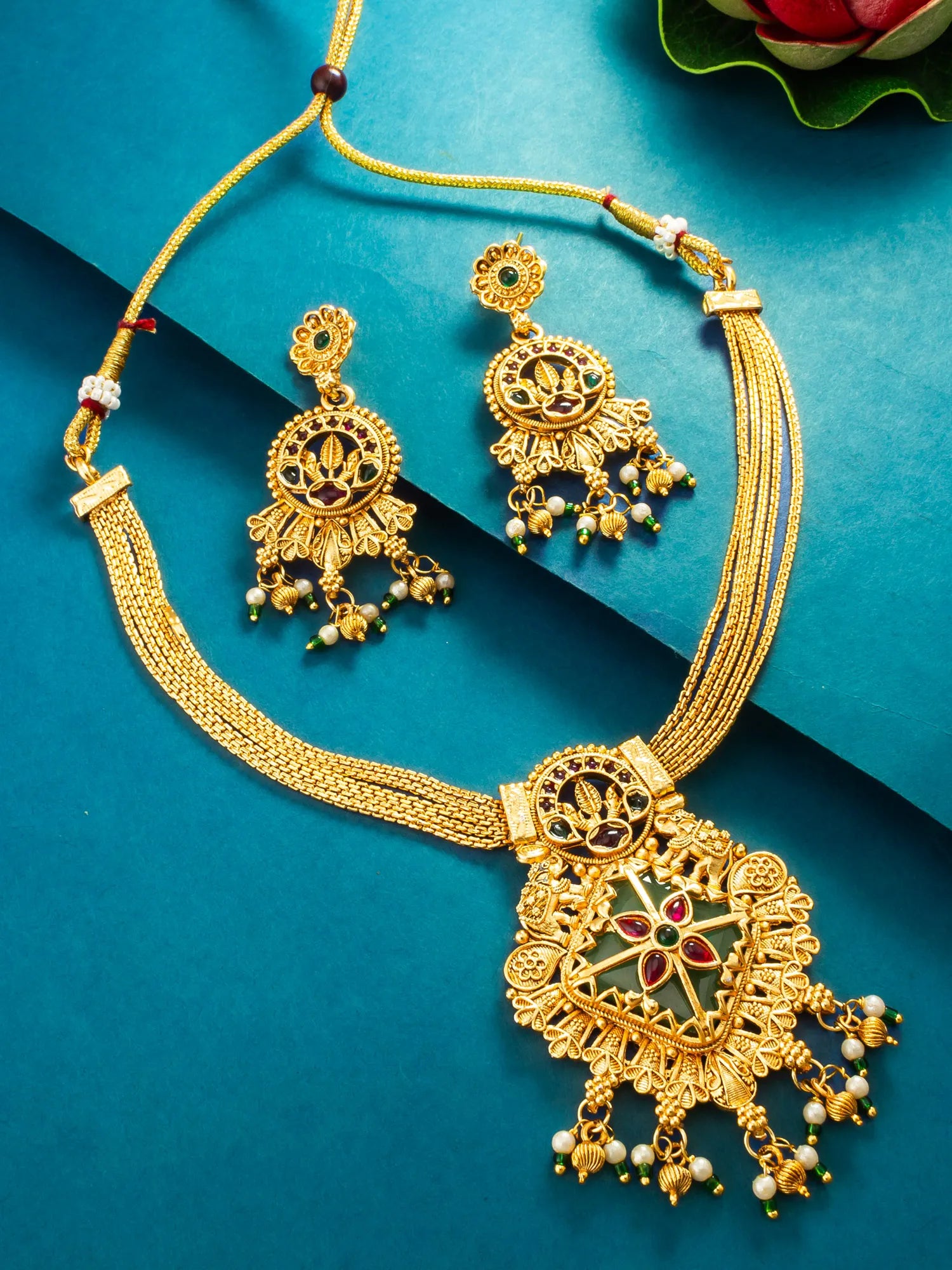 Gold-toned traditional jewelry set featuring a necklace and matching earrings with ornate filigree, red and green enamel details, and dangling pearl-like beads, arranged on a teal background.