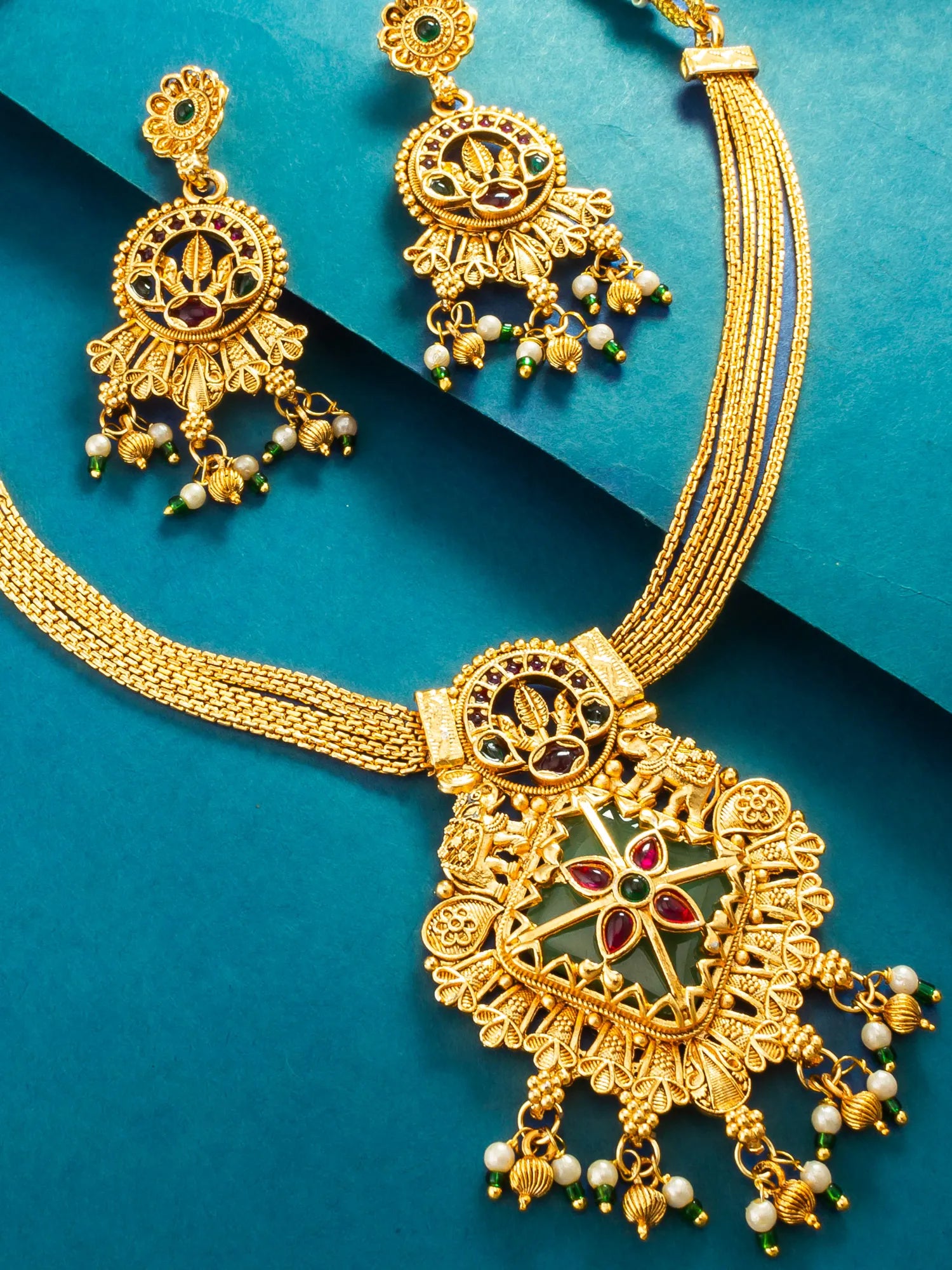 Gold-toned traditional Indian jewelry set featuring a necklace and matching earrings with intricate filigree work, red and green enamel details, and white pearl-like bead drops, arranged on a teal background.