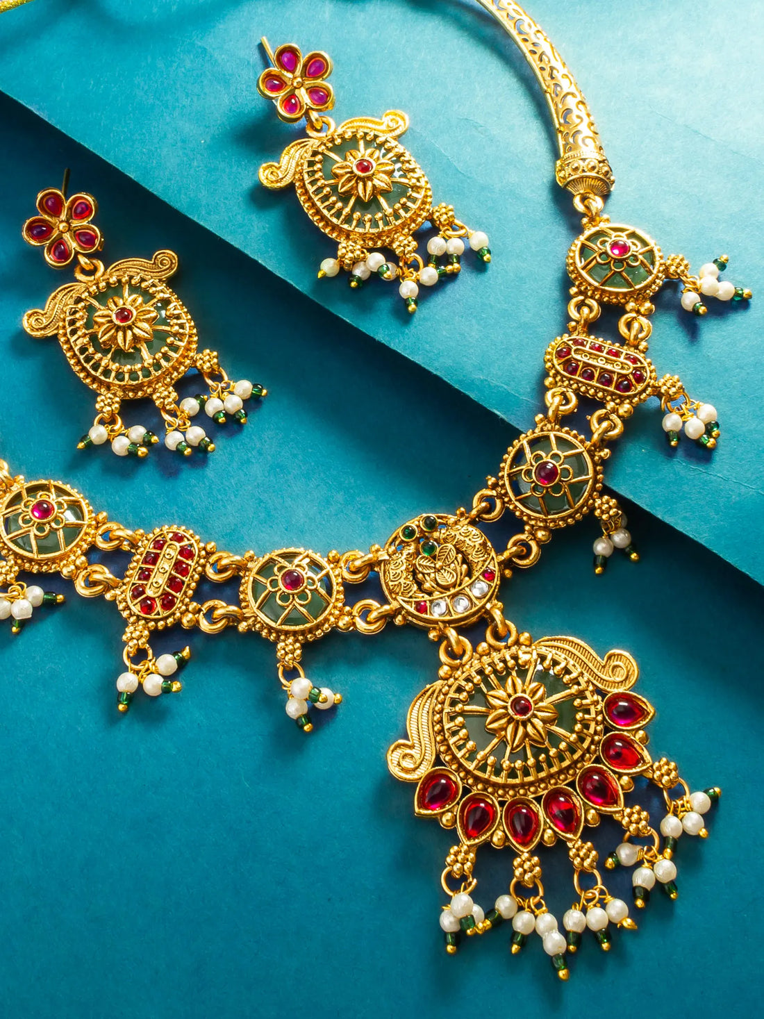 Gold-plated Indian jhumka necklace and matching earrings with red and green enamel details, set against a blue background.