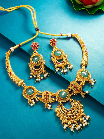 An ornate gold-toned traditional necklace and matching earrings set with green stone centers, red gem accents, and white bead drops, arranged on a blue background.