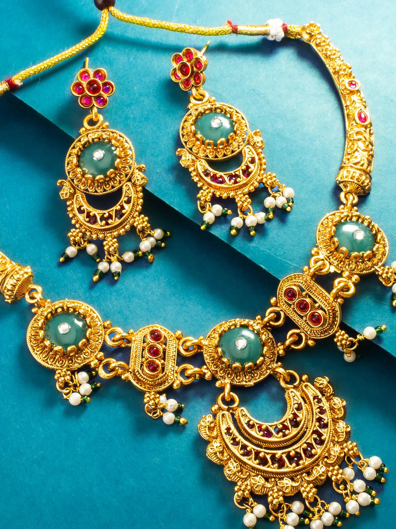 Gold-toned traditional jewelry set with a necklace and matching earrings, featuring green stone centers, red gemstone accents, and white pearl bead tassels, arranged on a blue background.