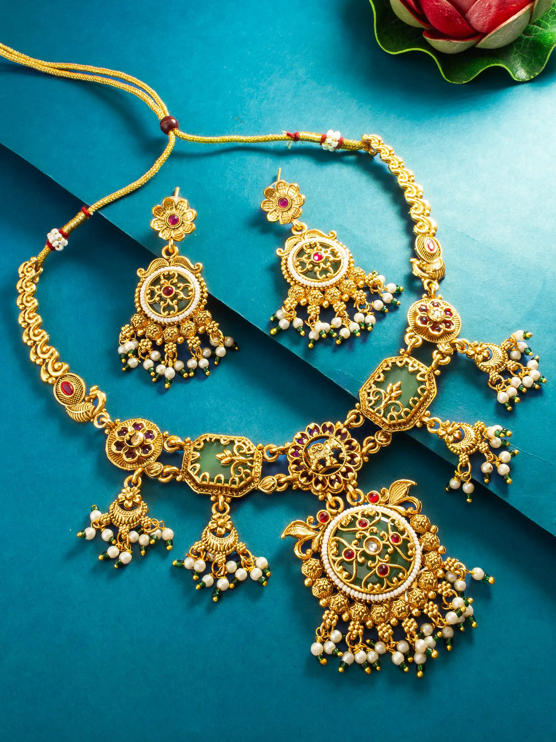 An ornate gold-toned traditional necklace and matching earrings with green enamel, red stone accents, and white pearl-like beads, arranged on a bright blue background.