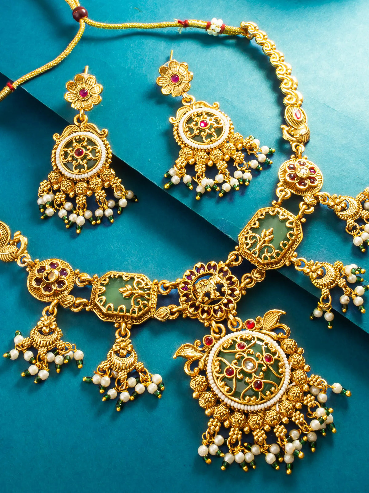 An ornate gold-toned jewelry set with matching necklace and earrings, featuring green enamel panels, white pearl-like beads, and small red gemstone accents, arranged on a teal background.
