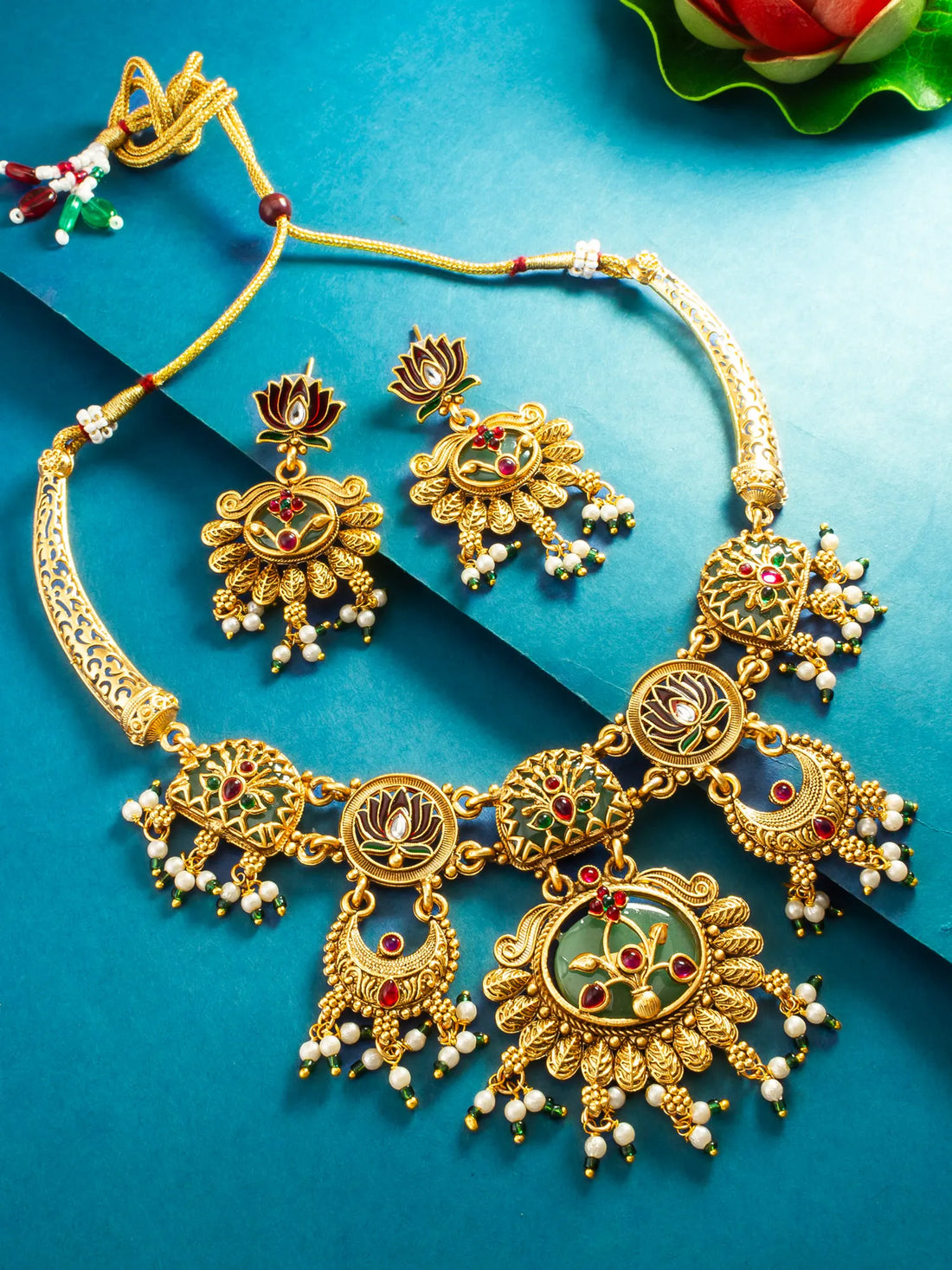 Gold-toned traditional Indian jewelry set featuring a necklace and matching earrings with floral motifs, red and green enamel details, and white pearl-like bead drops, arranged on a blue background.