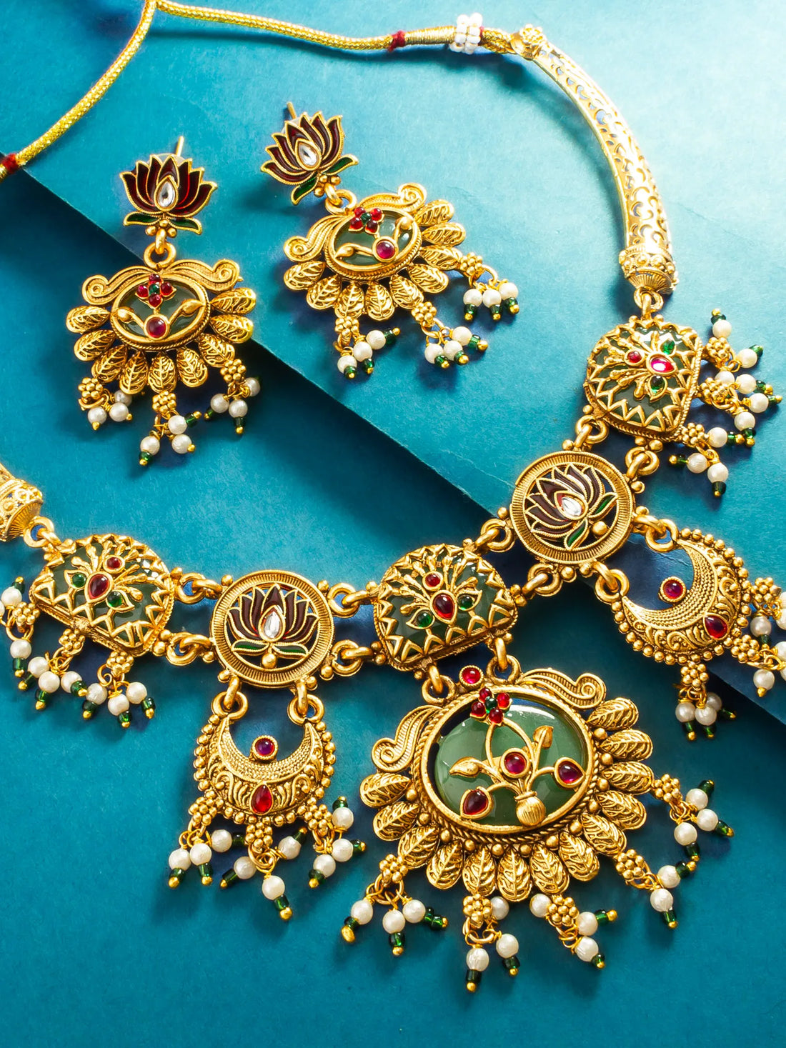 Gold-toned traditional Indian jewelry set featuring a necklace and matching earrings with floral motifs, red and green enamel details, and white pearl-like bead drops, arranged on a blue background.