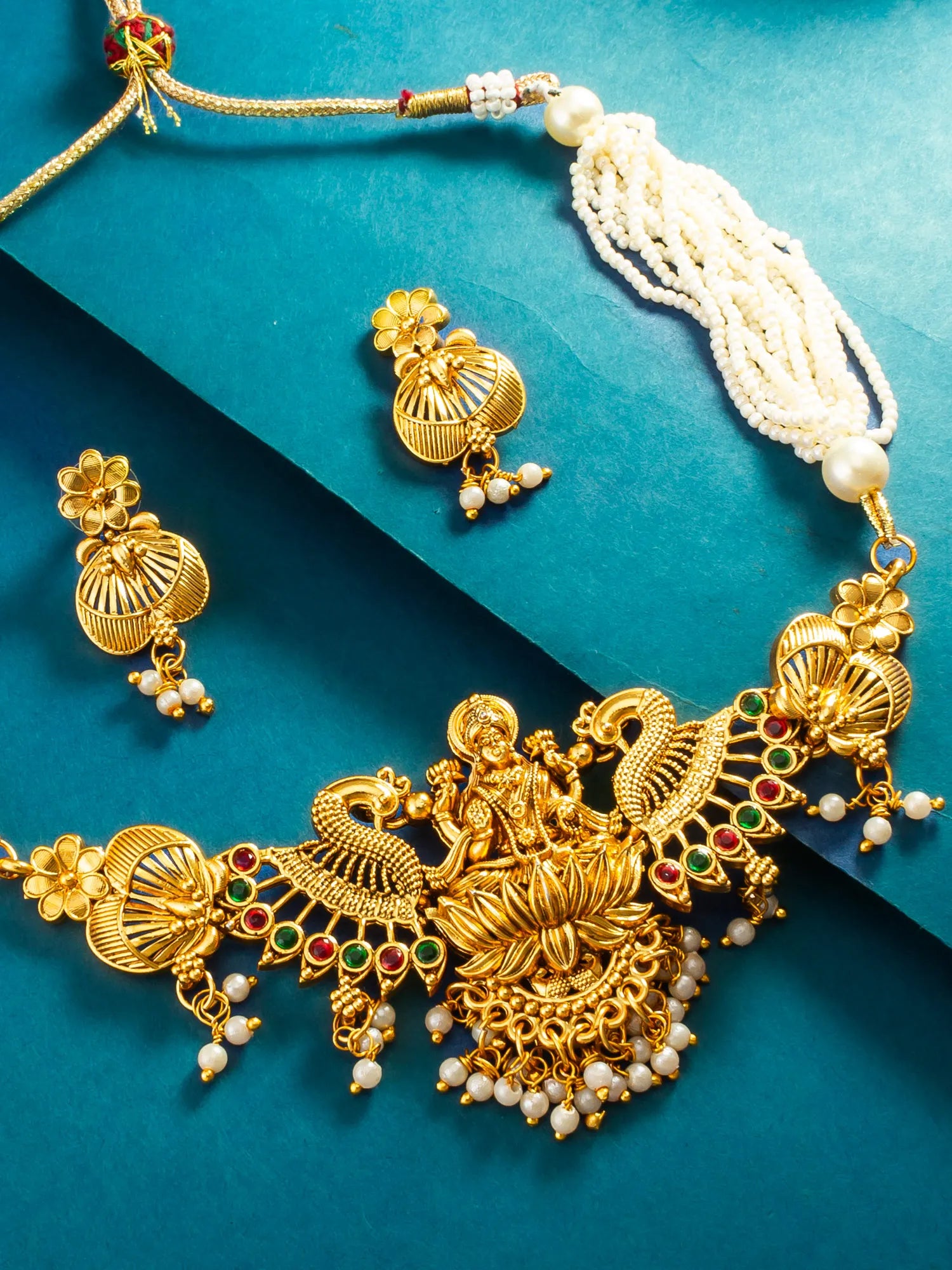 An ornate gold-toned traditional necklace set with matching earrings, featuring a central deity motif, peacock-like decorative elements, white pearl beads, and small red and green stone accents, arranged on a teal background.