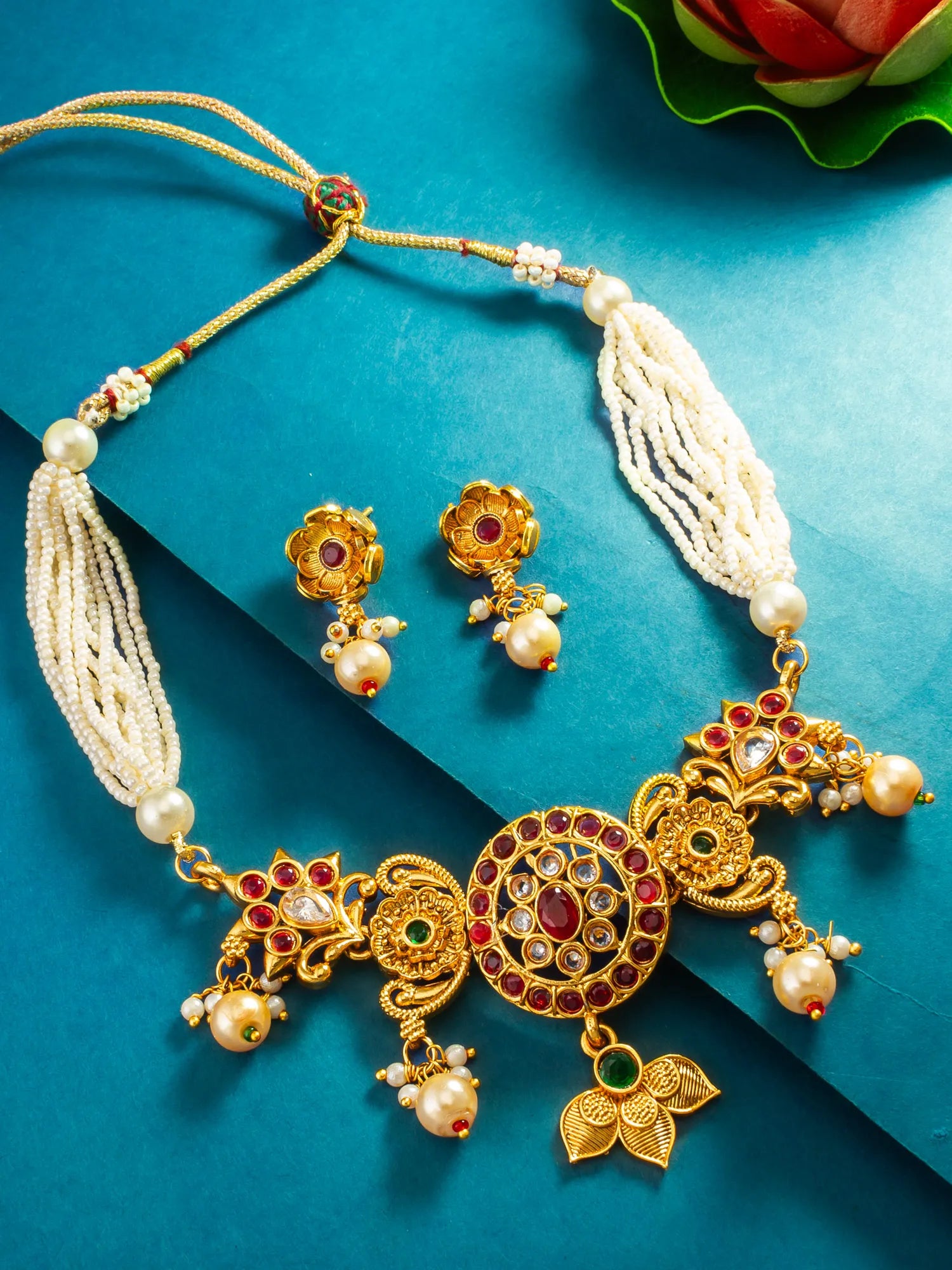 Gold-toned traditional Indian jewelry set featuring a necklace and matching earrings, embellished with white pearl-like beads, red and green stones, and floral motifs, arranged on a teal background.