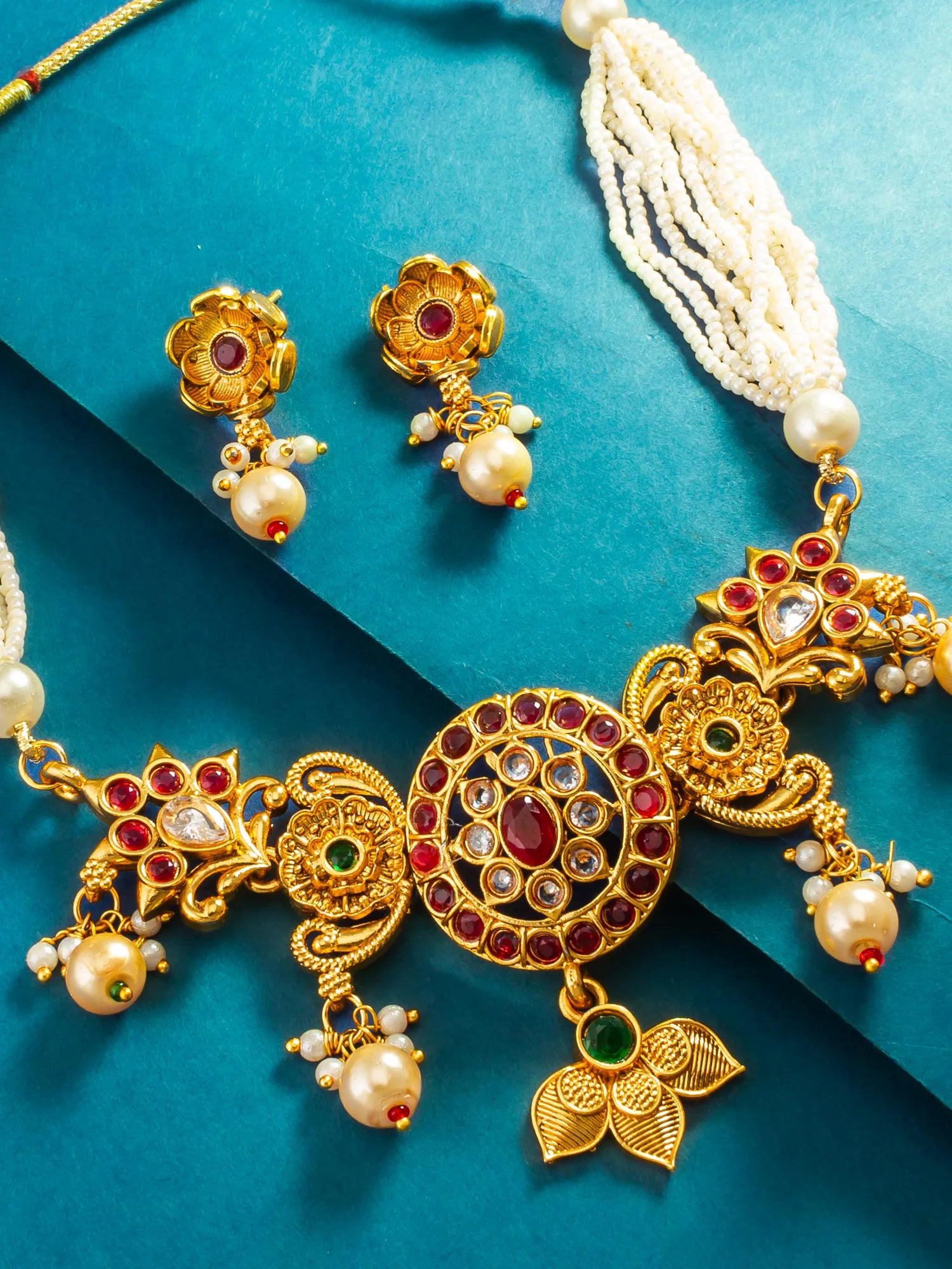 An ornate gold-toned traditional necklace set with matching earrings, decorated with red and green stones and white pearl-like beads, arranged on a teal background.