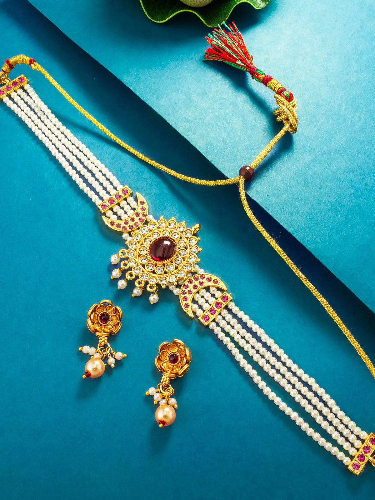 Gold-plated traditional Indian Kundan necklace with a central maroon stone, decorated with white pearl strands and red/green accents, shown with matching earrings on a teal background.
