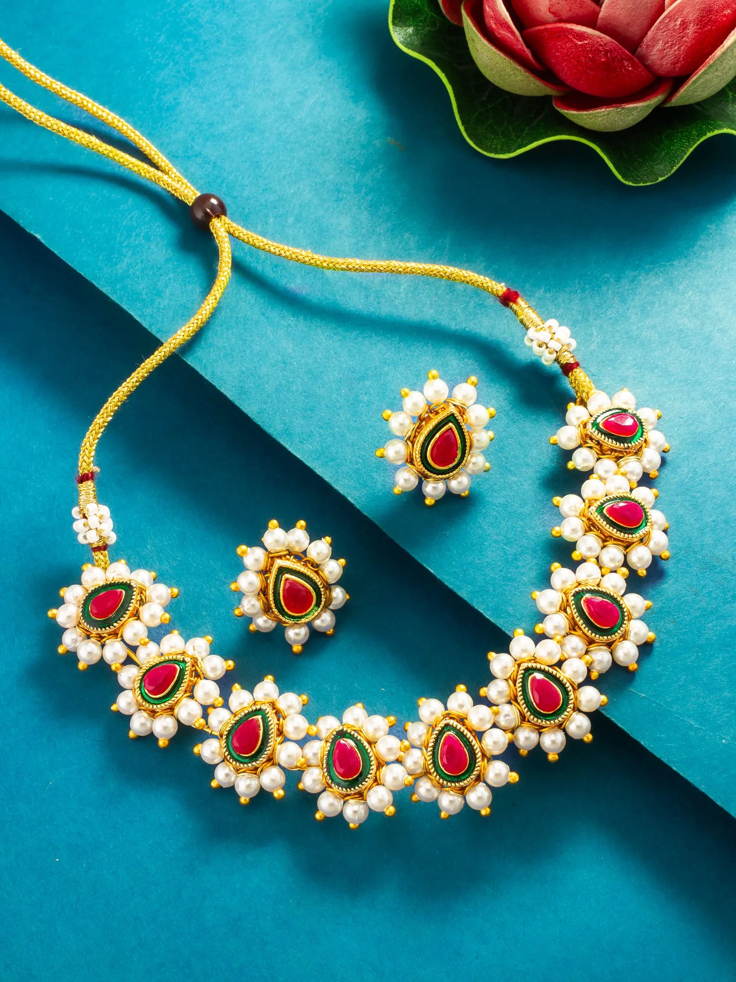 A traditional Indian jewelry set featuring a necklace and matching earrings with white pearl-like beads, red teardrop-shaped stones, and green and gold detailing, displayed on a textured blue background.