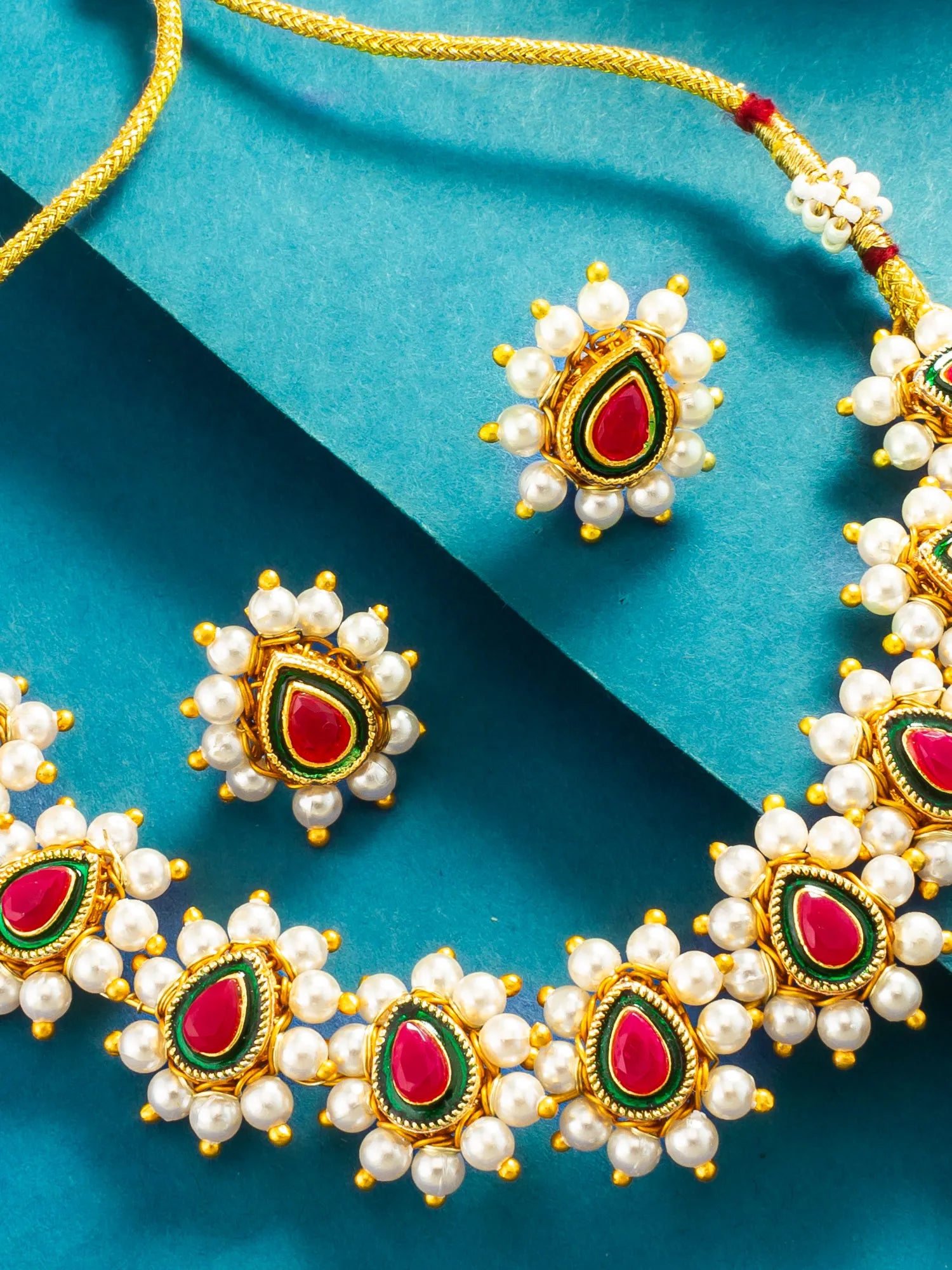 Gold-plated jewelry set featuring a necklace and matching earrings with white pearl-like beads, red teardrop-shaped stones, and green enamel accents, arranged on a teal background.